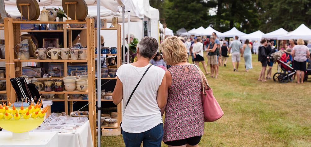 Two women seen from the back peruse a booth at the Muskoka Arts and Crafts Summer Show